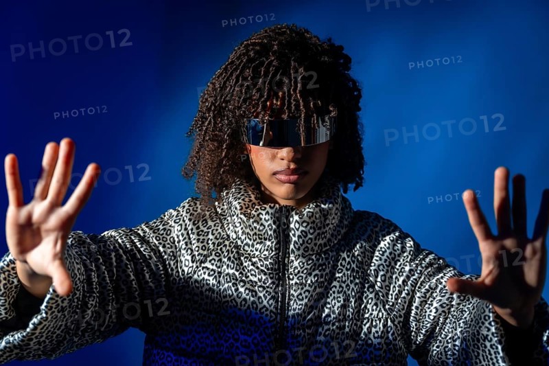 Young woman with futuristic visor and patterned jacket pushing virtual screen on blue background