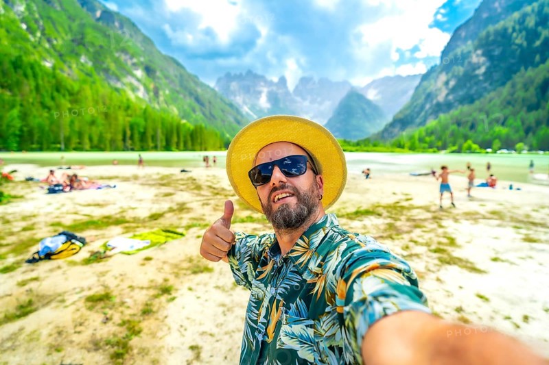 Tourist wearing sunglasses and straw hat gesturing thumbs up while taking a selfie at lago di landro with tourists enjoying summer in the dolomites, italy