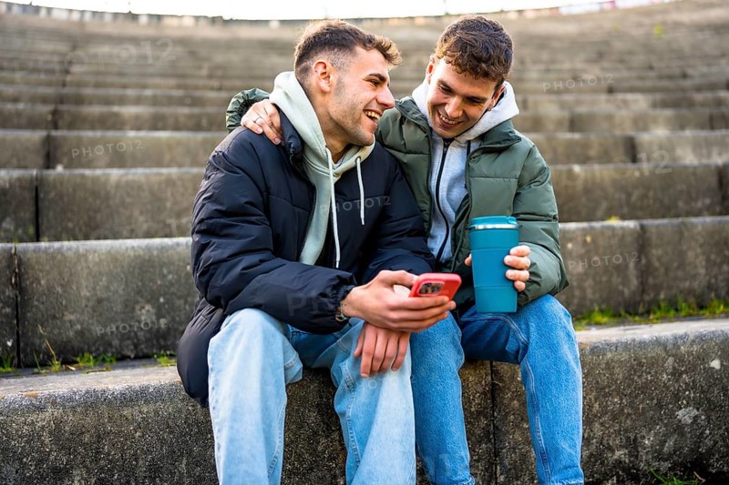 Two men are sitting on steps, enjoying a reusable coffee cup and sharing a laugh as they look at a smartphone together