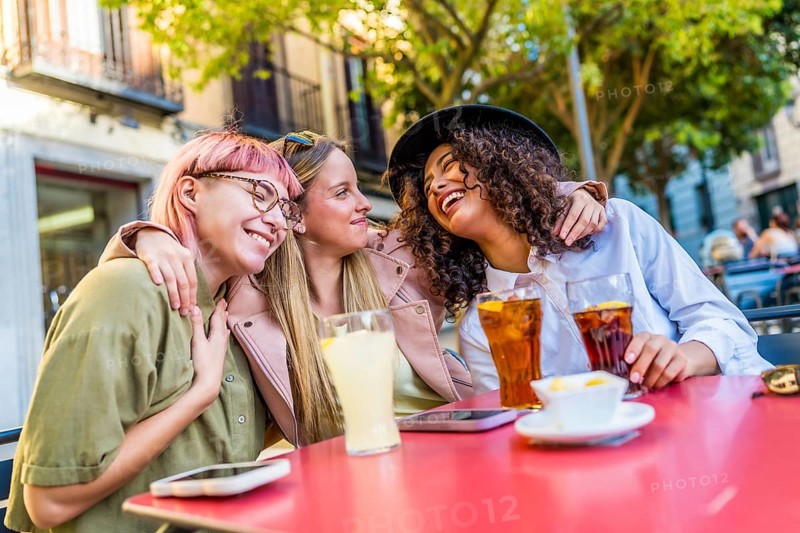 Diverse female modern friends hugging and smiling sitting in an outdoor bar terrace