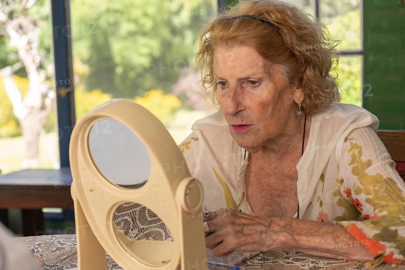 Senior woman applying makeup while gazing at her reflection in a double mirror on a table, illuminated by bright natural light streaming through a window in the background