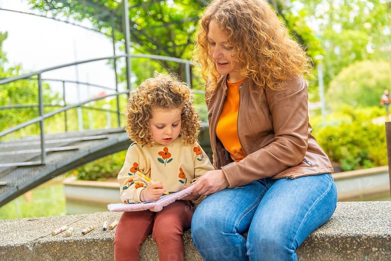 Blonde caucasian little girl learning to read on a park sitting with her mother
