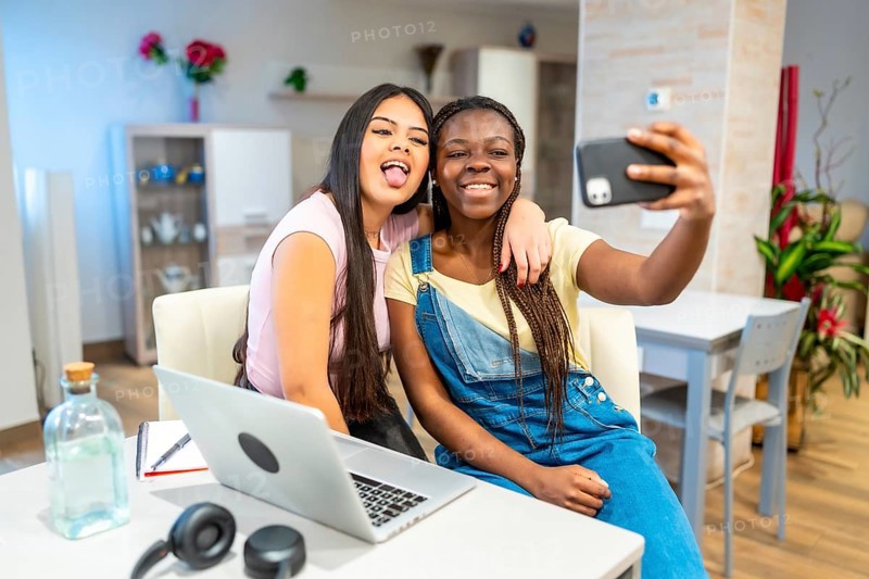 Two happy multi ethnic female students taking a selfie with a smartphone while studying at home