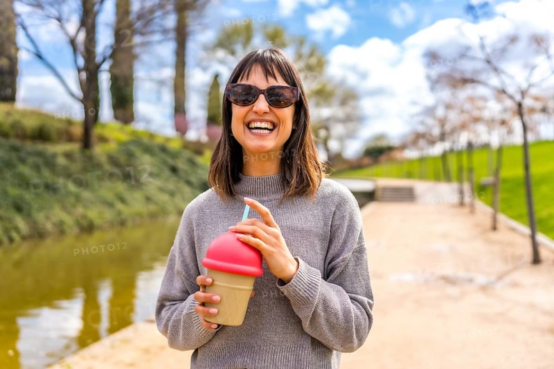 Young woman smiling and holding a reusable cup with a straw, enjoying a refreshing drink in a sunny park during springtime
