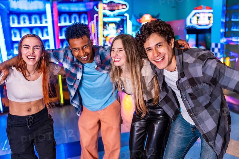 Four young cheerful multi ethnic friends are hugging each other, smiling and having fun together at the amusement arcade