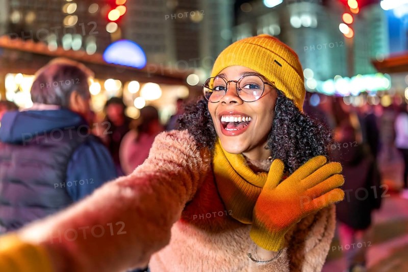 Cheerful young woman dressed in vibrant yellow winter clothes, capturing a joyful selfie amidst the festive lights of a bustling christmas market