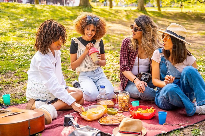 Four female friends are sitting on a blanket, eating snacks and drinking beverages during a picnic in a sunny urban park
