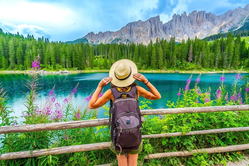 Female hiker with backpack admiring the turquoise waters of lake carezza and the latemar mountain range in the italian dolomites during summer