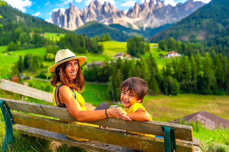 Mother and son sitting on a bench, admiring the breathtaking landscape of the italian dolomites near santa maddalena church in val di funes