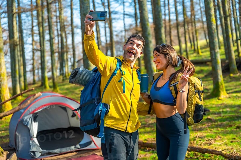 Happy couple of hikers taking a selfie with smartphone in a forest next to their tent during a camping trip