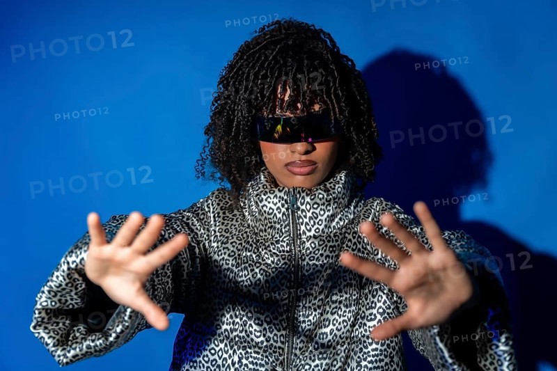 Young woman wearing futuristic visor and patterned jacket, gesturing with hands on blue background, immersed in metaverse gaming experience