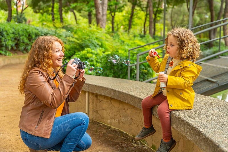 Mother taking portraits to her daughter blowing bubbles sitting on a park