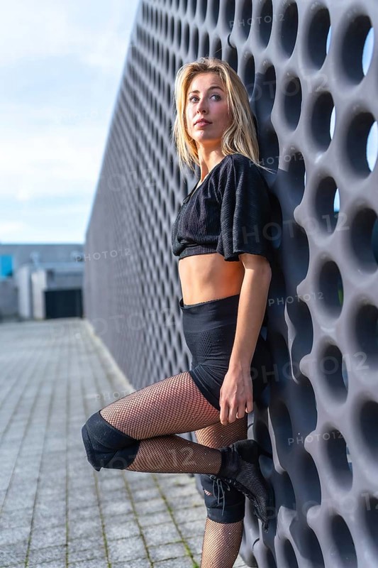 Young urban dancer striking a pose in high heels, knee pads, and fishnet stockings, exuding confidence and style against a city backdrop