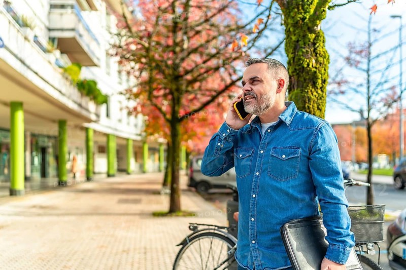 Mature man in denim clothes talking to the mobile and carrying laptop in the city in autumn