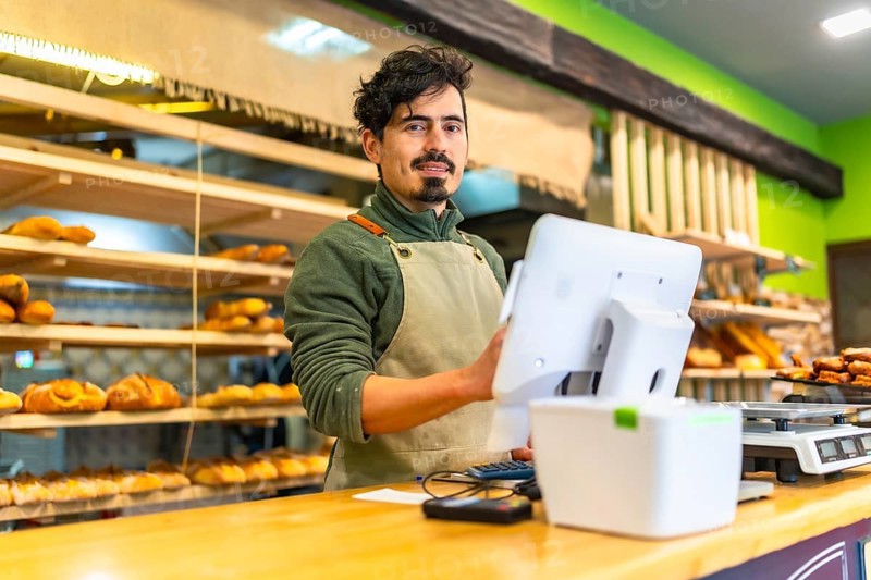 Latin friendly salesman in apron using computer stranding in the counter of a bakery shop