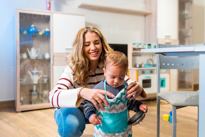 Happy mother helping her cute baby boy wearing a toy apron in the kitchen, playing and having fun together