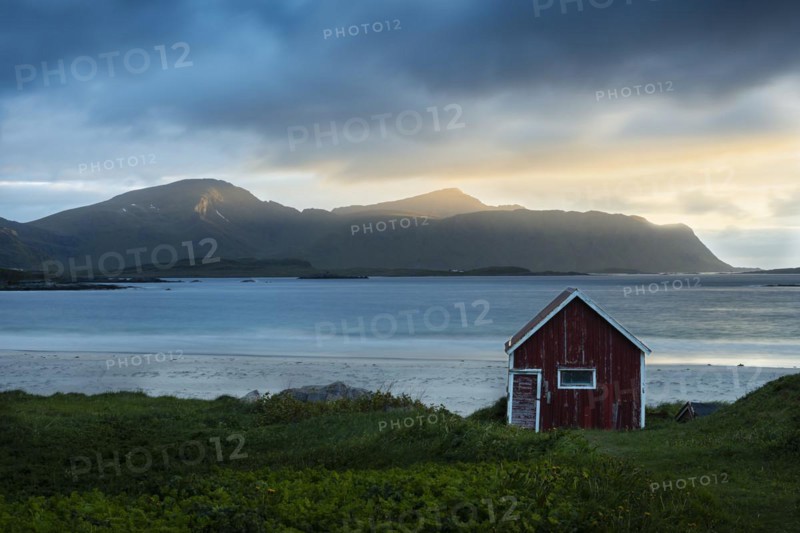 A red wooden hut on the sandy beach of Ramberg (Rambergstranda), with the sea behind it. At night at the time of the midnight sun. Clouds in the sky, the night sun shining from the side, mountain peaks in the sunlight. Early summer. Ramberg, Flakstadoya, Lofoten, Norway, Europe