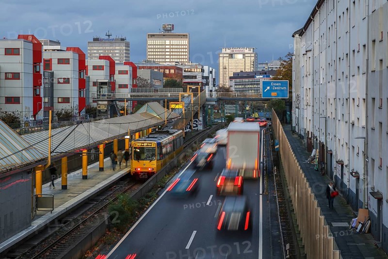Essen, North Rhine-Westphalia, Germany, motorway A40 in the city centre, exit Essen Zentrum, during dusk. Underground line U18 on the way to Berliner Platz stops at Savignystrasse underground station. On the right, residential buildings behind a noise barrier. A LANUV air monitoring station, Essen Kruppstrasse 117 (EKRU2), is located on the wall on the right under the lantern. Residents are affected by noise and exhaust fumes. Building with Postbank, Eon and Evonik lettering at the back, Europe