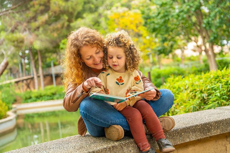 School girl sitting on the lap of her mother reading a book in a park