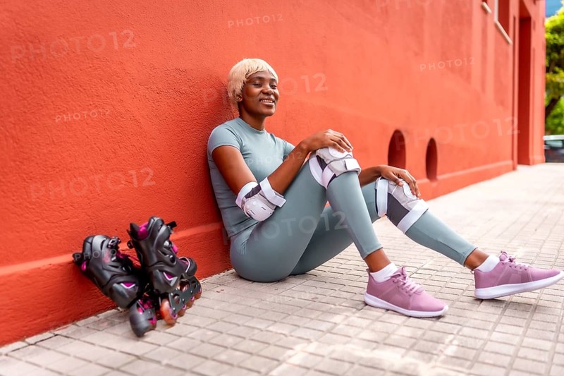 Beautiful african woman roller skating resting sitting on ground and leaning on red facade during the day