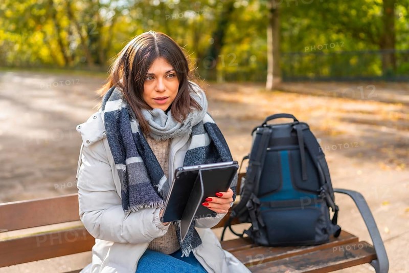 Woman with warm clothes working using digital tablet sitting on a park bench during sunny day of autumn
