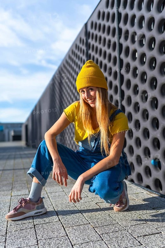 Smiling young woman dancer crouching in urban contest, wearing yellow t shirt, blue jeans and yellow beanie looking at camera