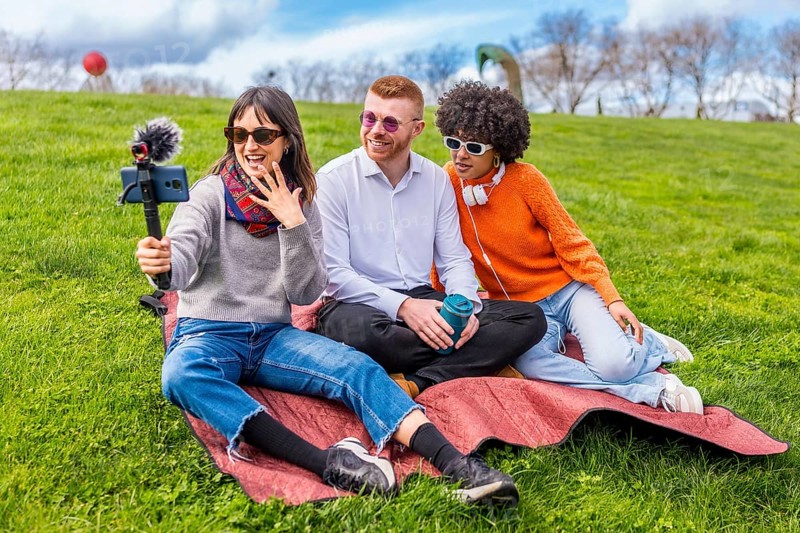 Three friends are recording a video blog in a park, using a smartphone with external microphone