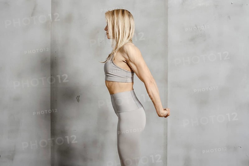 An athletically built woman stands in profile against the background of a concrete wall and stretches the muscles of her arms and back