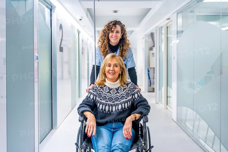 Happy nurse carrying mature female patient in wheelchair at the hospital corridor