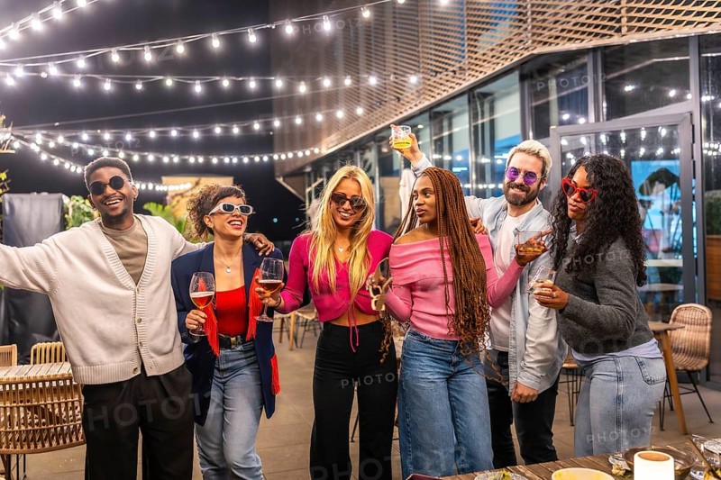 Group of young diverse multi-ethnic friends having fun at bar terrace decorated by lights at night