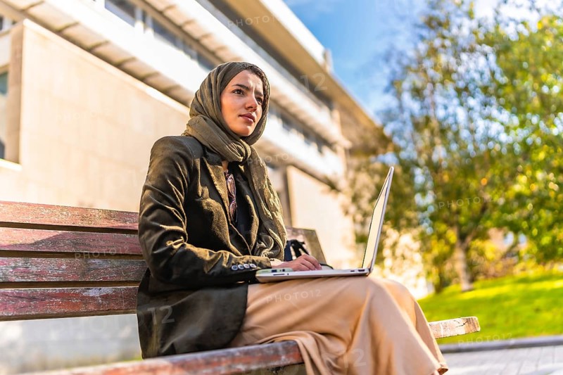 Side low angle view of a muslim businesswoman with headscarf working on remote sitting on park bench