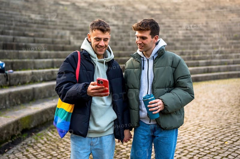 Happy gay couple holding hands, looking at smartphone and enjoying a reusable cup of coffee outdoors