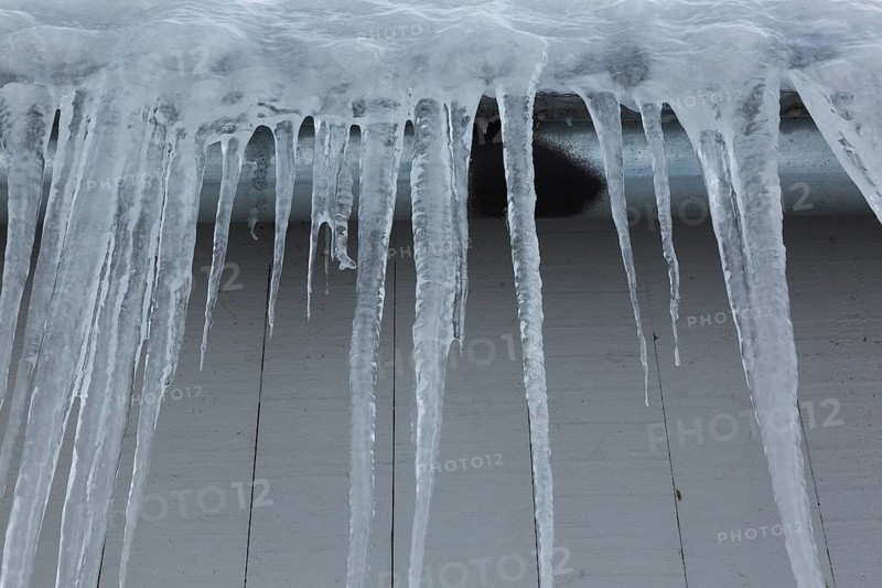 Long icicles and thick heavy ice hanging from roof edge of grey wood plank cladded building in winter, Quebec, Canada, North America