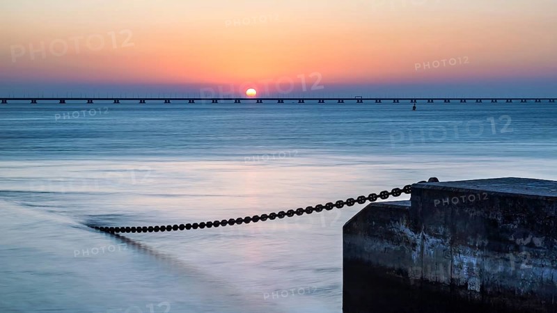 A serene sunset with the sun touching the horizon, beside a calm ocean and a pier extending into the distance, Lisbon