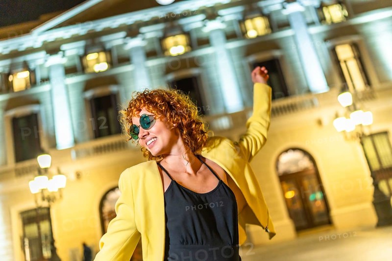 Happy woman in yellow jacket, black summer dress and sunglasses dancing alone in the city at night