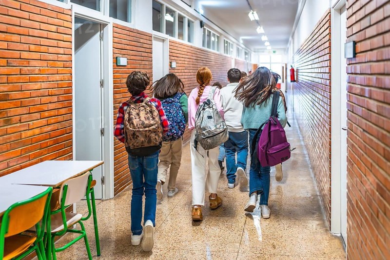 Rear view of elementary school students running down school hallway between classes, carrying backpacks