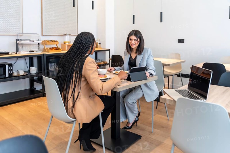 Two businesswomen enjoying breakfast and discussing work using a digital tablet in a modern hotel restaurant