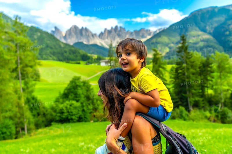 Mother carrying her son on her shoulders while enjoying the view of the odle mountains and the funes valley in the dolomites in summer