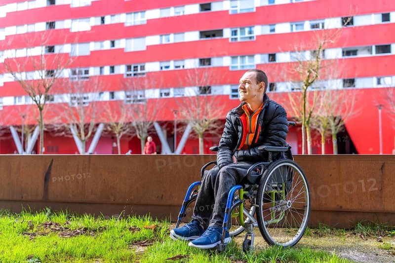 Young man sitting in wheelchair enjoying a sunny day in a modern urban park with a red building in the background