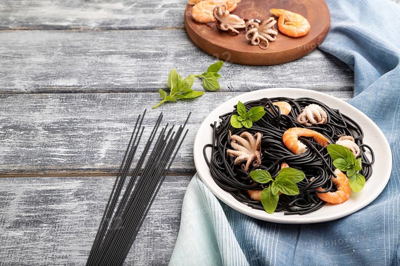 Black cuttlefish ink pasta with shrimps or prawns and small octopuses on gray wooden background and blue textile. Side view, copy space