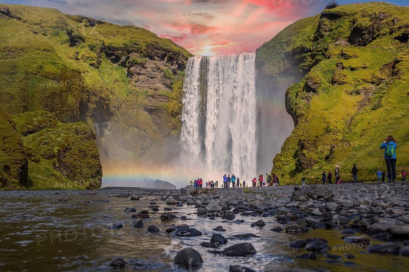Group of tourists looking at the beautiful rainbow at the impressive Skogarfoss waterfall in southern Iceland, photo taken in summer on a beautiful sunset