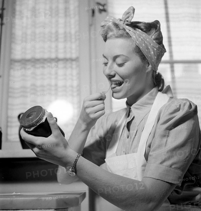 1940s practical tips. A young woman at home looks happy when licking the glue side of a label to put on the glass jar of the jam she has made herself. She is in the kitchen dressed in an apron and has her hair set up in a typical way. The year is 1940 and the world war II is going on. Food and other necessities are rationed, therefore an own supply of food was neccesary. Photo Kristoffersson 158-1