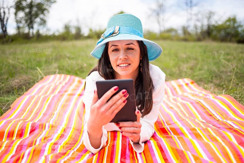 Attractive smiling young woman dressed casually uses e-book/tablet lying down on a colourful blanket in the park. Modern lifestyle using portable mobile devices everywhere people go. Reading e-mail
