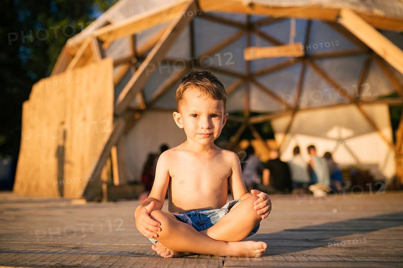 Theme is yoga and children. Caucasian Boy child sitting barefoot cross-legged in lotus position on wooden floor the background of gathering place of people. In summer sunset. Dressed in denim shorts