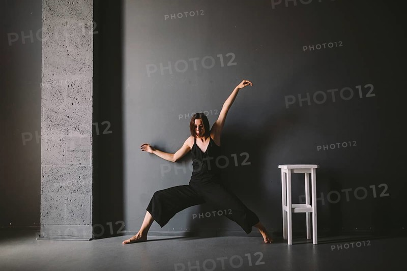 Social theme of female loneliness is pain suffering. Abastration of male domestic violence. Young beautiful Caucasian woman in black clothes, strange pose, white chair big dark room. Fear and despair
