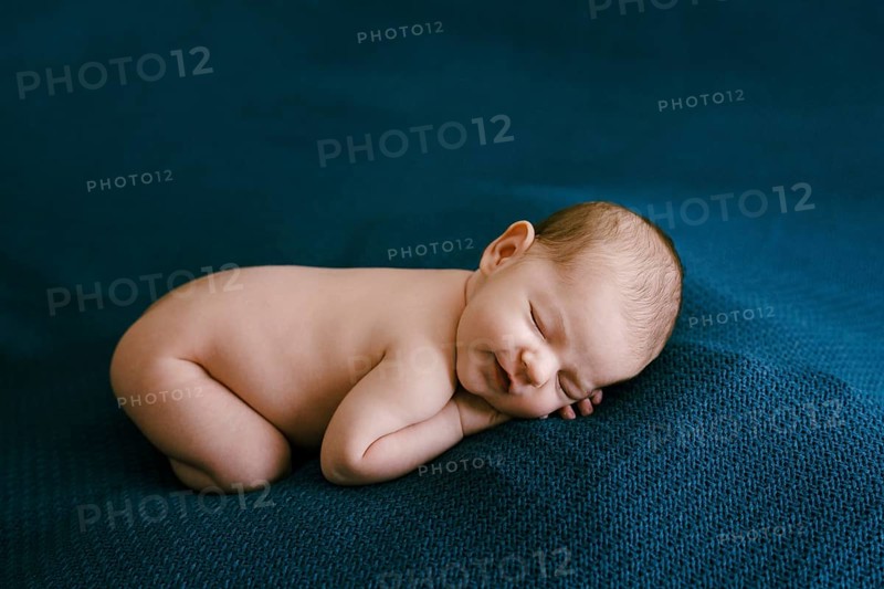 Naked newborn sleeping on a blue blanket with arms and legs tucked in.