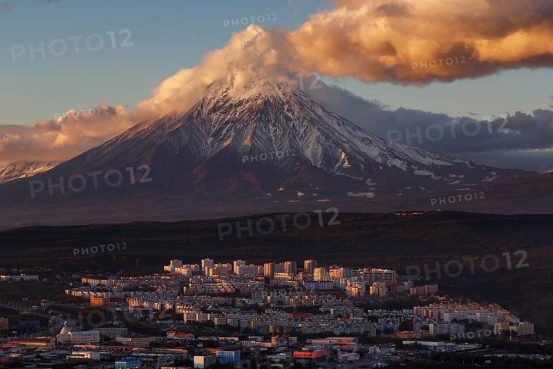 Evening colorful top view of townscape of Petropavlovsk-Kamchatsky City at sunset on background clouds drifting across blue sky, cone of active Koryak Volcano. Kamchatka Peninsula, Russian Far East