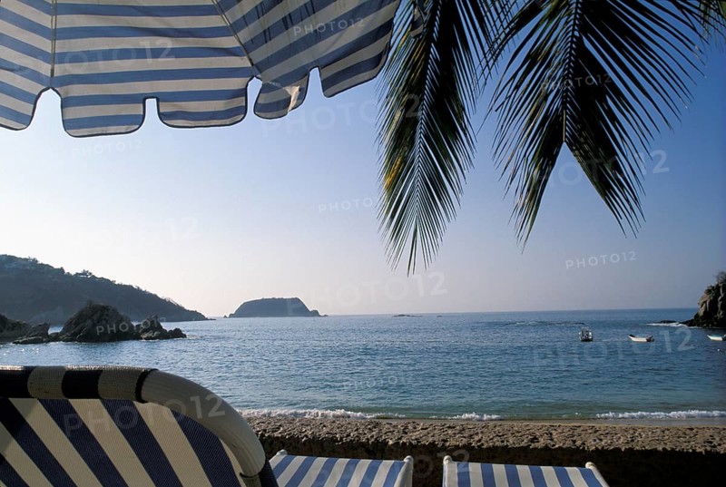 A tranquil beach scene with palm leaves, a striped umbrella, and a view of the ocean and distant island, in the coast of Mexico