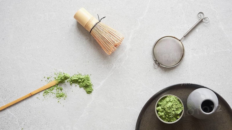 Matcha green tea powder and utensils for drink preparation: traditional bamboo spoon and whisk. Top view on gray stone table