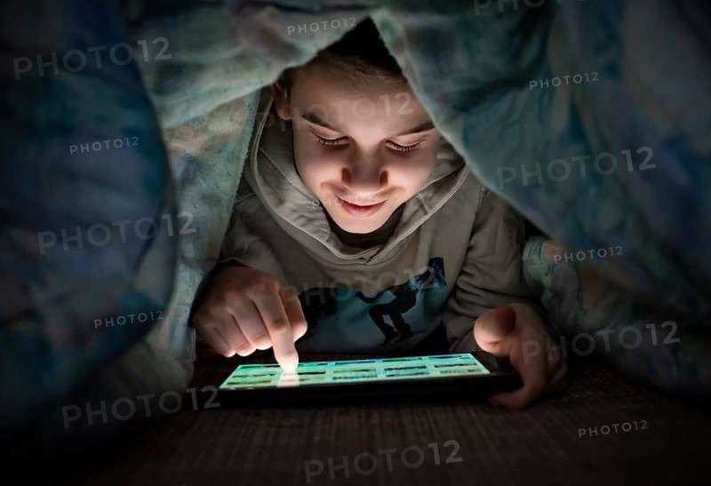 Child watching his tablet in the bed. Illuminated child face from device screen. Smiling Boy under the covers hold a tablet. Night time